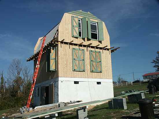 east end of the gambrel roofed cabin with plywood siding, green shutters and the roof sheathed